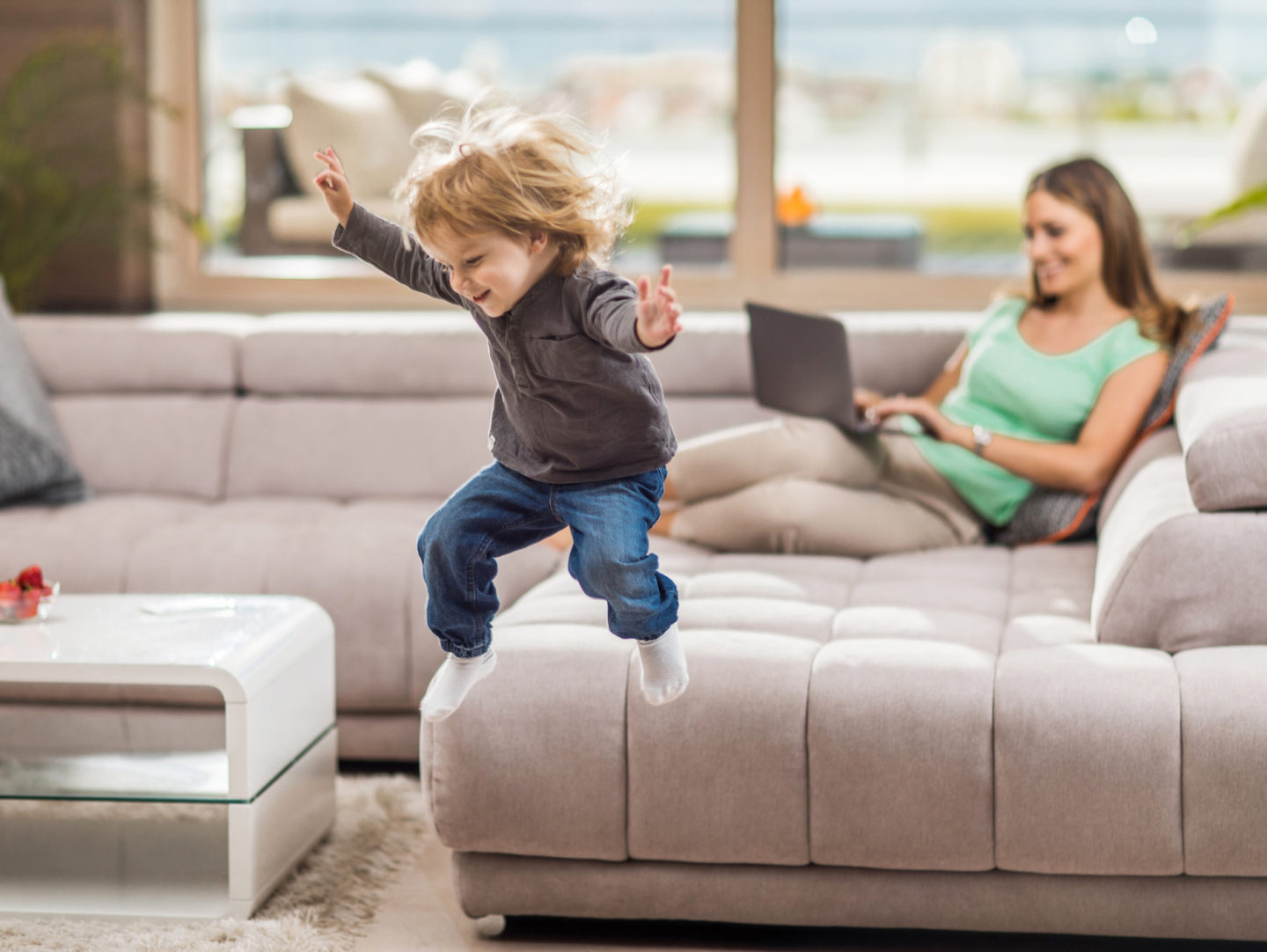 Glad pojke hoppar från soffan hemma. Hans mamma använder en laptop i bakgrunden.